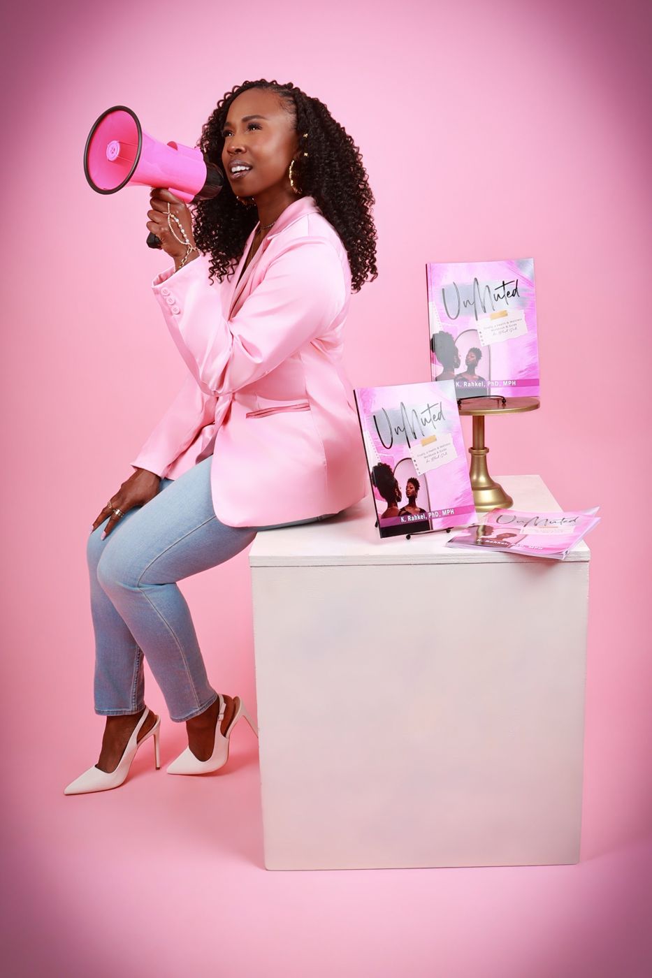 Dr. K. Rahkel in a pink blazer speaking into a pink megaphone beside copies of UnMuted